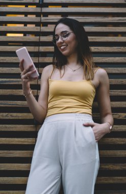 Neşeli bir kadın cep telefonuyla internete göz gezdirirken akıllı telefonuyla modern binanın ahşap duvarının yanında dikiliyordu.