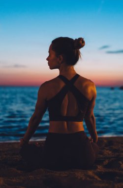 Gün batımında kumlu deniz kıyısında oturmuş dinlenirken ve gözlerini kaçırırken meditasyon yapan zayıf genç bir kadının arka görüntüsü.