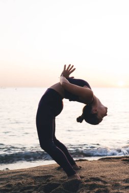 Spor giyimli, esnek genç bir bayanın güneş batarken kumlu sahilde Urdhva Dhanurasana 'da yoga egzersizi yaparken tam boy yan görüntüsü.