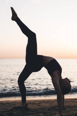 Spor giyim kuşağında yoga yapan genç yogi kadın Eka Pada Urdhva Dhanurasana 'yı gün batımında boş plajda seyrediyor.