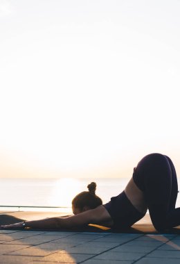 Sıska spor elbiseli genç bir kadın güneş batarken deniz kenarında halı üstünde yoga yaparken poz veriyor.