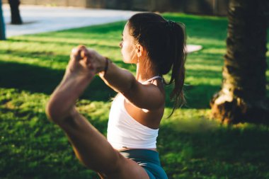 Spor kıyafetleri içinde, gün batımında yoga yapan genç ve esnek bir kadın. Temiz havada yeşil çimlerin üzerinde yatıyor.