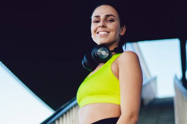 Aktif giyinmiş, modern kulaklıklı, neşeli beyaz kadın portresi. 20 'li yaşlarda, sağlıklı yaşam tarzını korumak için dışarıda merdivenlerin yanında duran mutlu kadın koşucu.