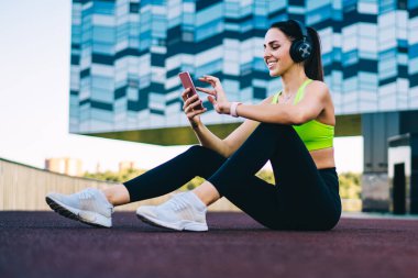 Şehir merkezinde oturan ve spor salonundaki insanlarla sabah sporu hakkında mesajlaşan mutlu formda bir antrenör. Kulaklıklı neşeli bir kadın, pozitif sesli kitap dinliyor.
