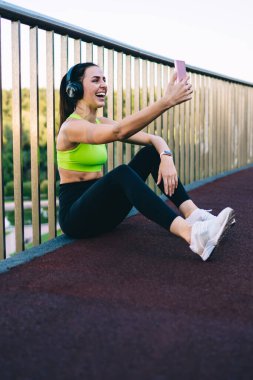 Sıska, genç, gülen spor kadın spor kıyafetleri ve kulaklıklı spor ayakkabılar metal eskrim ve akıllı telefon kullanırken Frendc 'le görüntülü sohbet ederken.