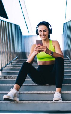 Gülümseyen zayıf bayan koşucu, akıllı telefon uygulamasında sabah egzersizinden memnun. Güzel kadın koşucu, merdivenlerde modern cep telefonu kullanarak sosyal ağlarda eğitim almanın içeriğini paylaşıyor.