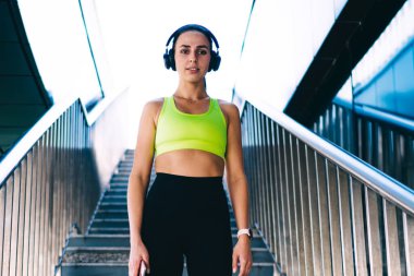Aktif haldeki zayıf beyaz kadın koşucu merdivenlerde ayakta durup kulaklıkla müzik dinliyor, mükemmel vücut yapısı olan bayan sporcu sağlıklı yaşam tarzını korumak için ses aksesuarı kullanıyor.
