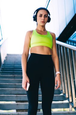 Modern binanın merdivenlerinde dururken kulaklıkla müzik dinleyen kararlı, zayıf kadın koşucunun düşük açılı görüntüsü.