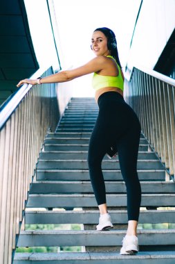 Üst katta yürüyen şık spor kıyafetleri ve beyaz spor ayakkabıları giyen, kameraya bakarken kulaklıkla ve akıllı telefonuyla müzik dinleyen genç, pozitif açılı genç bir bayan.