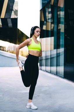 Genç sporcu kadın vücut esnekliği antrenmanı sırasında dış kardiyo egzersizi yapıyor. Elektronik bluetooth kulaklıklı aktif güçlü kadın ses kitabı dinliyor ve bacak kaslarını esnetiyor.