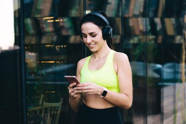 Neşeli bin yıllık kadın dışarıda spor yapıyor. Mesajlaşma ve çevrimiçi mesajlaşma için cep telefonu aygıtı kullanıyor. Renkli eşofmanlı kadın elektronik bluetooth ekipmanlarıyla radyo dinliyor.