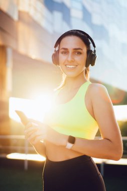 Genç, modaya uygun spor giyim sektöründe atletik bir kadın akıllı telefon kullanıyor ve arka planda durup cam bina ve kameraya bakarken çalma listesinden şarkılar dinliyor.