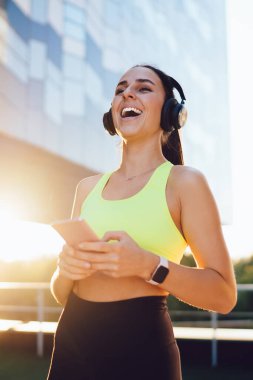 Elektronik kulaklıklı dişlek bayan sporcu pozitif ses setini dinliyor ve güneşli kentselliğe gülüyor, akıllı telefon cihazlı neşeli bir kız sabah antrenmanı için seviniyor.