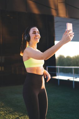 Aktif giyinmiş beyaz kadın koşucu güneşli bir sabahta akıllı telefon kamerasına poz veriyor kulaklıklı, gülümseyen bir spor kadını telefonla sohbet ediyor.