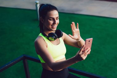 Spor kıyafetleri ve beyaz spor ayakkabılar giyen neşeli, zayıf bayan koşuculardan metal eskrimin yanında duran ve akıllı telefondan video çağrısı yapan.