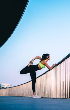 Formda bayan koşucunun şık spor kıyafetleri ve spor ayakkabıları giyip bacağını ısıtırken metal korkuluklara yaslanıp başka yere bakarken ve kulaklıkla müzik dinlerken yan görüntüsü.