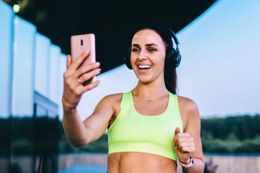 Spor giyim kuşağında tatmin olmuş, gülümseyen ve sohbet eden bir bayan koşucu. Şehirdeki bir binanın yanında durmuş, kablosuz kulaklıkla müzik dinliyor.