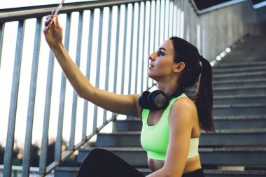 Spor sutyen ve taytlı atletik bir kadının metal merdivenlerde oturması ve sokak eğitiminden sonra dinlenirken akıllı telefondan selfie çekmesi.