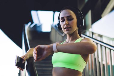 Modern binanın yanındaki merdivenlerde dururken trabzanlara yaslanan ve fitness takip cihazını kontrol eden modaya uygun aktif giyimli genç sporcu bayandan.