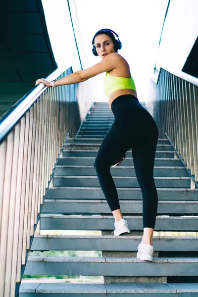 Açık havada vücut kaslarını çalıştırmak için kameraya bakan ince kadın sporcunun tam boy portresi bluetooth kulaklıklı spor hipster kız şehir merkezinde gürültü önleyici poz verdiği için poz veriyor.