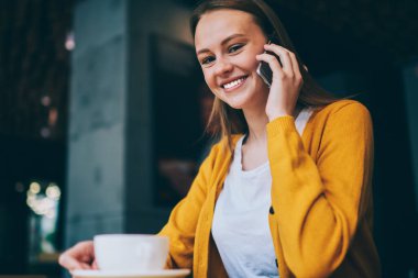 Kafenin içindeki akıllı telefon konuşmalarında kameraya gülen neşeli yeniyetme kızın portresi çay saatinde kafeteryada arkadaşça telefon görüşmesi yapmaktan keyif alan sıradan giyinmiş mutlu bir kadın.