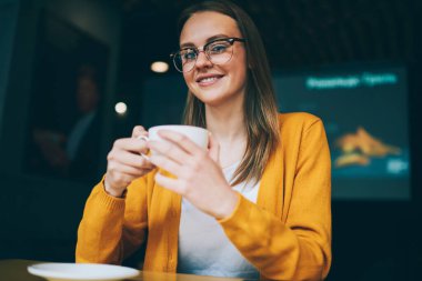 Kafeteryadaki kahve molası sırasında kameraya bakıp göz koruması sağlayan şık gözlüklü pozitif kadın portresi, kafein bardağı tutan başarılı hippi kız.