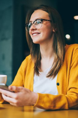 Gözlüklü, gözlüklü, elinde akıllı telefon aleti ve elinde fincanı olan mutlu bir kadın. Kafeteryada iletişim kurmak için cep telefonu bağlantısını kullanırken yüzünü çeviren pozitif hippi bir kız.