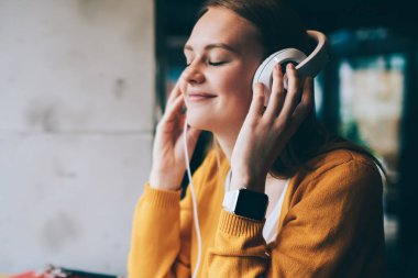 Elektronik kulaklıkla romantik müzik listesinden hoşlanan pozitif kadın meloman, kulaklıklı mutlu hippi kız modern laptopla kafede oturmuş romantik sesli kitap dinliyor.