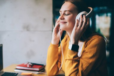 Elektronik kulaklıkla romantik müzik listesinden hoşlanan pozitif kadın meloman, kulaklıklı mutlu hippi kız modern laptopla kafede oturmuş romantik sesli kitap dinliyor.