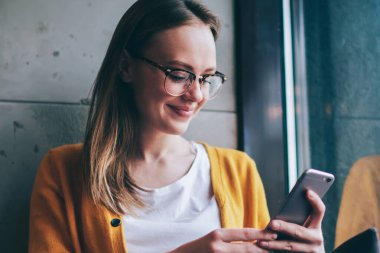Görüş düzeltmesi için modaya uygun gözlüklü mutlu kadın internet sayfasında olumlu haberler okurken akıllı telefondan internete göz atıyor, başarılı kadın blogcu pencere kenarında oturuyor ve SMS iletisi iletisi bağlıyor