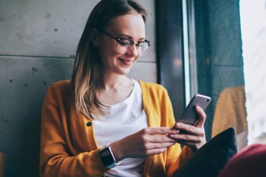 Görüş düzeltmesi için modaya uygun gözlüklü mutlu kadın internet sayfasında olumlu haberler okurken akıllı telefondan internete göz atıyor, başarılı kadın blogcu pencere kenarında oturuyor ve SMS iletisi iletisi bağlıyor