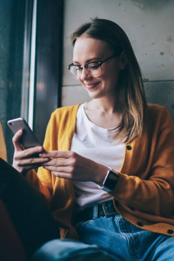 Görüş düzeltmesi için modaya uygun gözlüklü mutlu kadın internet sayfasında olumlu haberler okurken akıllı telefondan internete göz atıyor, başarılı kadın blogcu pencere kenarında oturuyor ve SMS iletisi iletisi bağlıyor