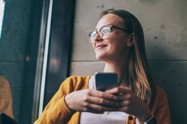 Gözlüklü mutlu kadın pencerenin kenarında oturuyor ve içeride dostça bir telefon bekliyor, elinde akıllı telefon tutan neşeli hippi kız ve kablosuz internete bağlanmış güncelleme bildirimi