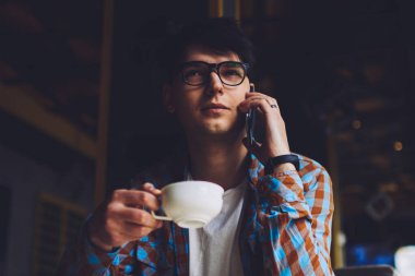 Erkek müdür cep telefonuyla konuşuyor ve lezzetli kahve içerken bazı konuları tartışıyor. Gözlüklü bir iş adamı kafeteryanın içinde telefonla iletişim kuruyor ve sıcak içeceğin tadını çıkarıyor.