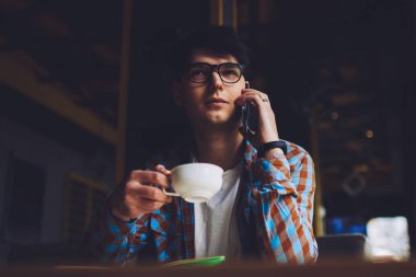 Erkek müdür cep telefonuyla konuşuyor ve lezzetli kahve içerken bazı konuları tartışıyor. Gözlüklü bir iş adamı kafeteryanın içinde telefonla iletişim kuruyor ve sıcak içeceğin tadını çıkarıyor.