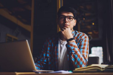 Optik gözlüklü zeki öğrencilerin portresi kameraya bakıyor kitaplara bakıyor ve hızlı internet bağlantısı olan modern bilgisayarlara bakıyor. Kafenin içinde serbest çalışan erkek metin yazarı.