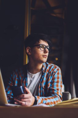 Moda gözlüklü, modern akıllı telefonu elinde tutan düşünceli bilişim uzmanı dizüstü bilgisayarın başında oturup pencereden dışarı bakıyor. Kafenin içinde serbest çalışan düşünceli bir metin yazarı.