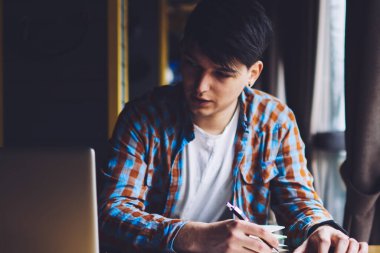 Zeki hippi, dizüstü bilgisayara bakıyor ve not defterine bilgi yazıyor. Kafede oturmuş internetten ilginç bir Webinar izlerken not alan dalgın genç adam.