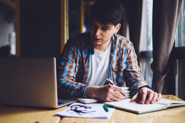 Zeki hippi, dizüstü bilgisayara bakıyor ve not defterine bilgi yazıyor. Kafede oturmuş internetten ilginç bir Webinar izlerken not alan dalgın genç adam.