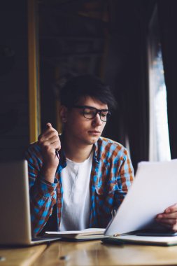 Zeki öğrenci kağıt üzerinde yazılı metin bilgilerini okuyor. Diz üstü bilgisayar ve kitaplarla şık bir kahve dükkanında oturuyor. Hipster adam elinde kalem tutuyor ve derslere bakıyor.