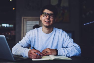 Daha iyi görebilmek için gözlüklü yakışıklı bir öğrencinin portresi. Elinde kalemle masada oturuyor ve bazı metin bilgileri yazıyor. Laptop bilgisayarında okuyan olumlu zeki bir genç adam.