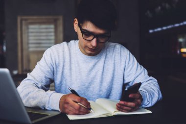 Gözlüklü bir öğrenci dizüstü bilgisayarla ahşap masada otururken akıllı telefondan not defterine metni yeniden yazıyor. Elinde telefon olan hippi adam bilgileri not alıyor.