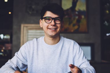 Daha iyi bir vizyon için gözlük takan mutlu yetenekli öğrenci modern telefonu elinde tutuyor ve kafenin iç kısımlarında masabaşına bakarken bir yandan da kompozisyon yazıyor.