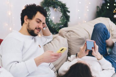 Beyaz erkek milenyumu, kış tatillerinde elinde cep telefonuyla şenlikli bir dairede kanepeye uzanmış kız arkadaşı cep telefonuyla e-posta gönderirken yakışıklı bir adam kameraya bakıyor.