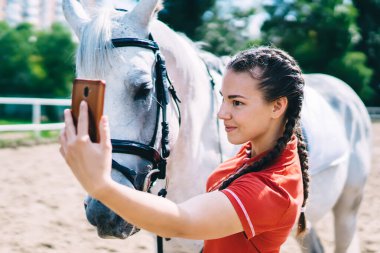 Beyaz kadın jokey otlakta at sırtında boş zamanın keyfini çıkarıyor ve sosyal ağlarla medya içeriğini paylaşmak için fotoğraf çekiyor, milenyum arkadaş canlısı kısrakla selfie vlog yapıyor