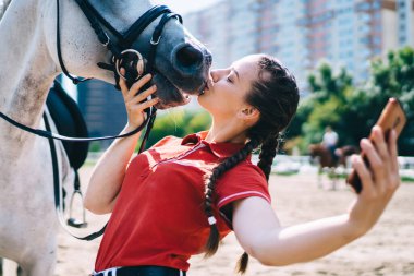 Beyaz kadın jokey otlak kulübünde safkan kısrakla öpüşüyor ve sosyal ağlarla medya içeriğini paylaşmak için selfie çekiyor, bin yıllık arkadaş canlısı aygırla fotoğraf çekiyor.