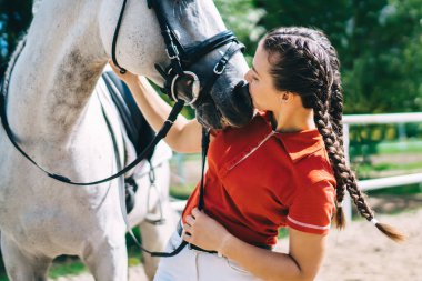 Güzel esmer profesyonel kadın jokey aygır ve öpüşen atın yanında duruyor, kadın binici çiftlikte tatildeyken hayvan arkadaşına bakmaya ve onunla ilgilenmeye hazırlanıyor.