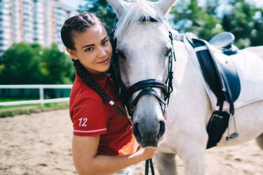 Özel bir üniforma giymiş, beyaz şampiyon bir atla kameraya bakıp boş zamanların tadını çıkaran, binicilik sporu konsepti olan kaygısız genç bir kadının yarım boy portresi.