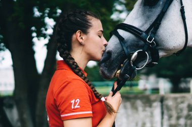 Hafta sonu at arkadaşlarıyla antrenman yapan kaygısız kadın jokeyin, safkan aygır kısrağa aşık beyaz jokeyin çiftlikte eğlenirken hayvanları öptüğünün yan görüntüsü.