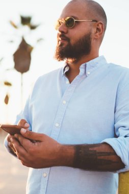 Sohbet için yenilikçi adam dövme kafe ayakta açık havada gün batımında seçtiğiniz için cep telefonu uygulamasını kullanarak el ile gezici 4 g bağlantısı kullanarak akıllı telefon tutarak yakışıklı sakallı erkek